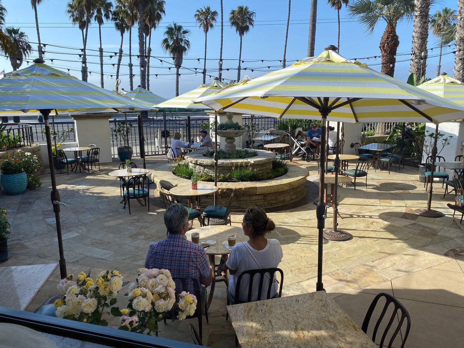 Sunny café patio with striped umbrellas, palm trees, ocean views, and people enjoying drinks near yellow roses.