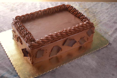 A rectangular cake covered in smooth chocolate frosting with a piped chocolate border around the top edge. The sides are decorated with large dark chocolate shards, and the cake is displayed on a gold cardboard base.
